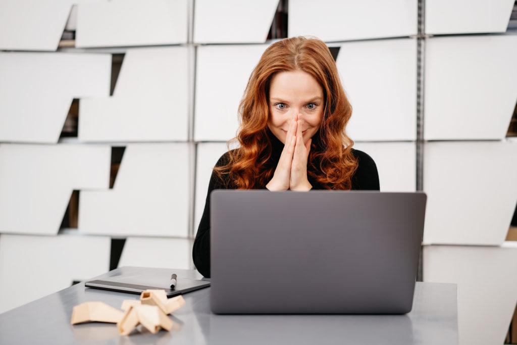 Excited young businesswoman looking at her laptop with a wide eyed look of anticipation and amazement as she sits at a table in a modern office