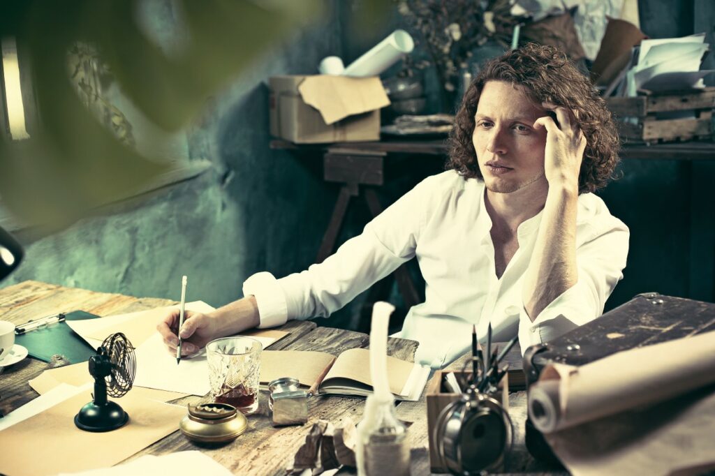 A moody looking man with long curly hair, staring into space as if thinking, while holding a pen to a piece of parchment paper.